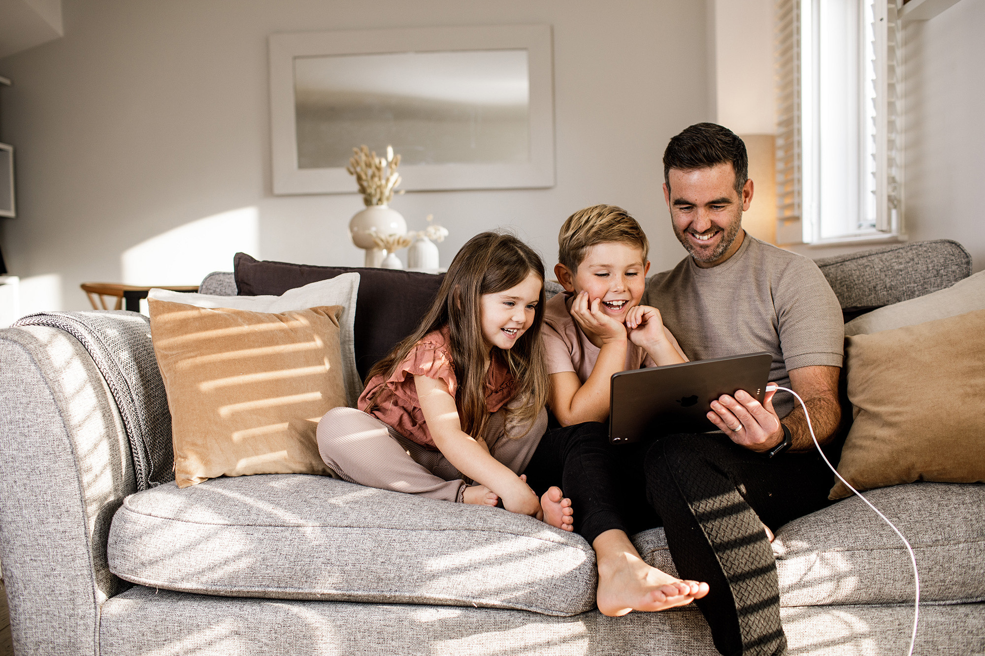 A father and his children enjoy watching a tablet whilst sitting on a comfortable sofa.