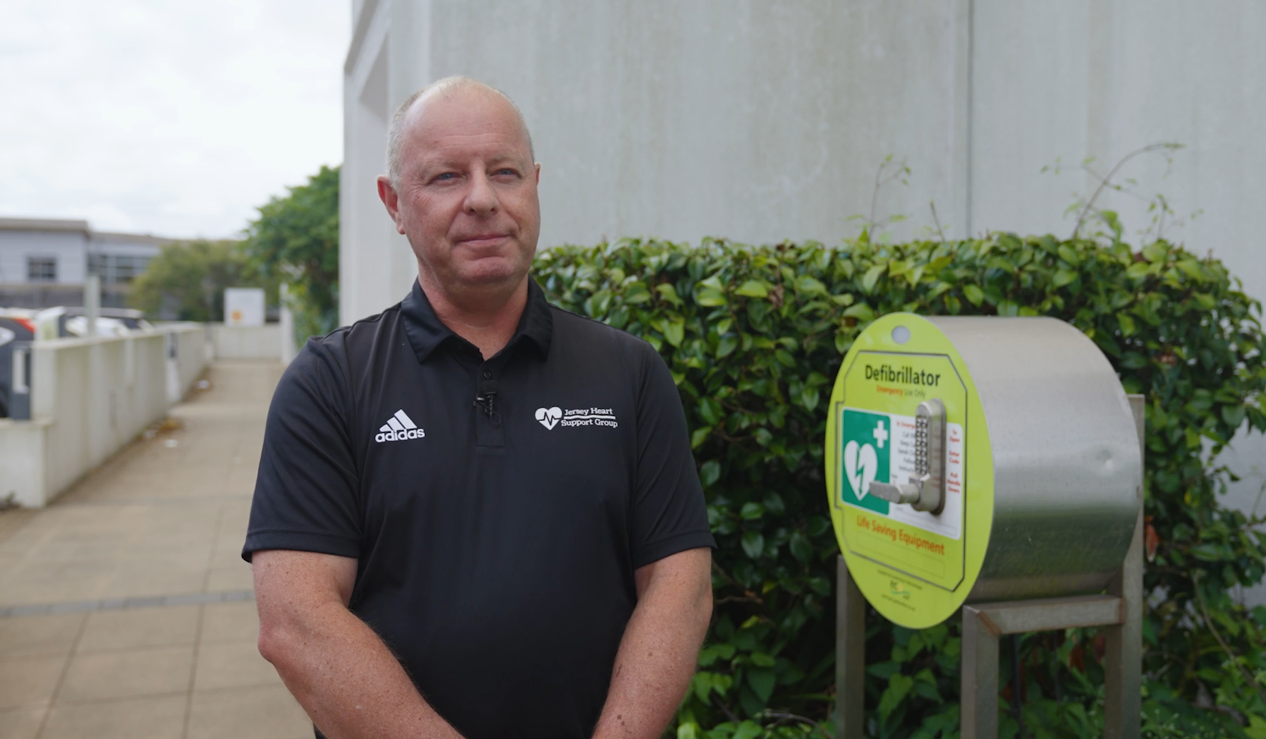 A person in a blue t-shirt standing outside a building, next to a defibullator