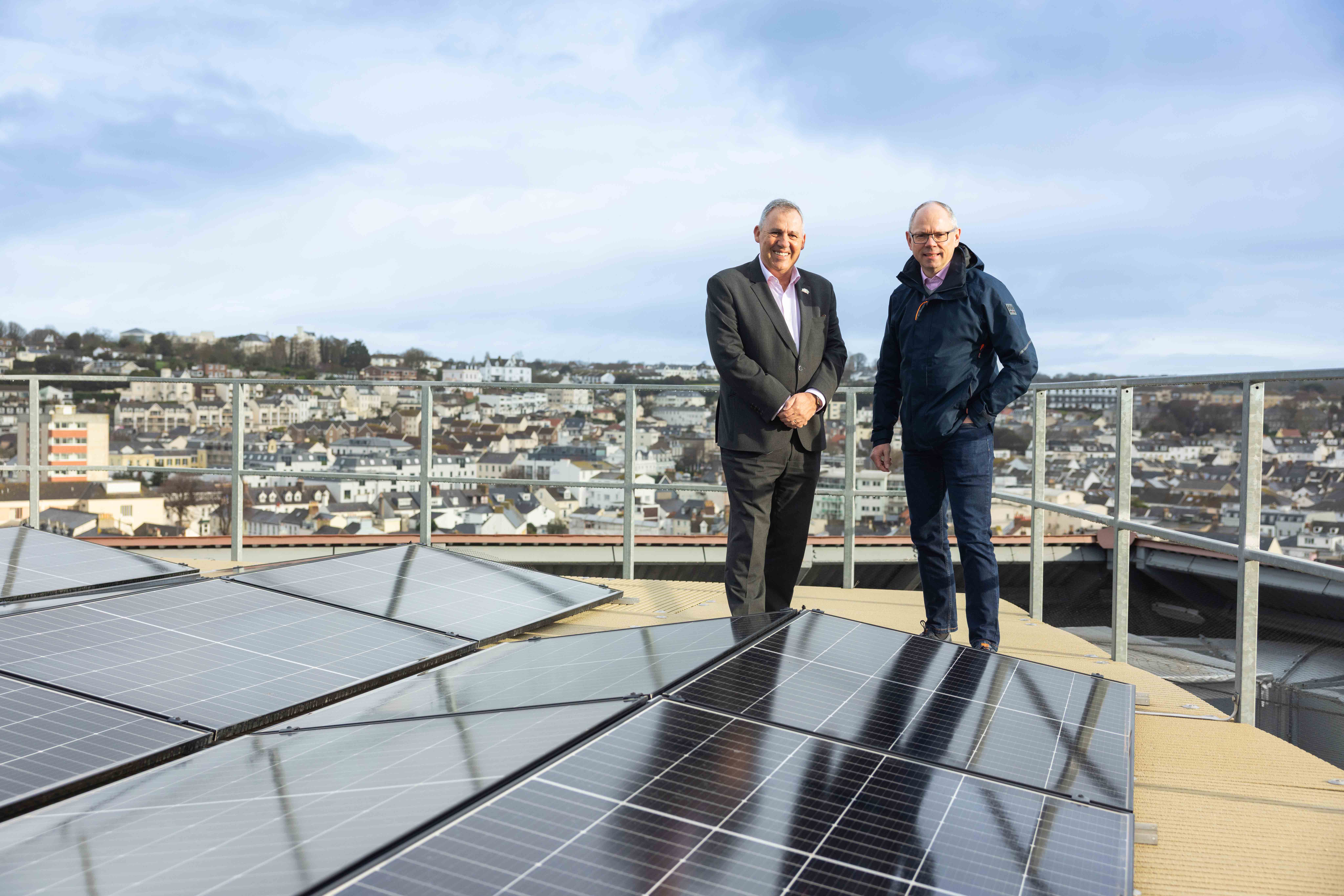 Two people dressed in suits standing on a rooftop next to solar PV panels. There are town buildings in the background.