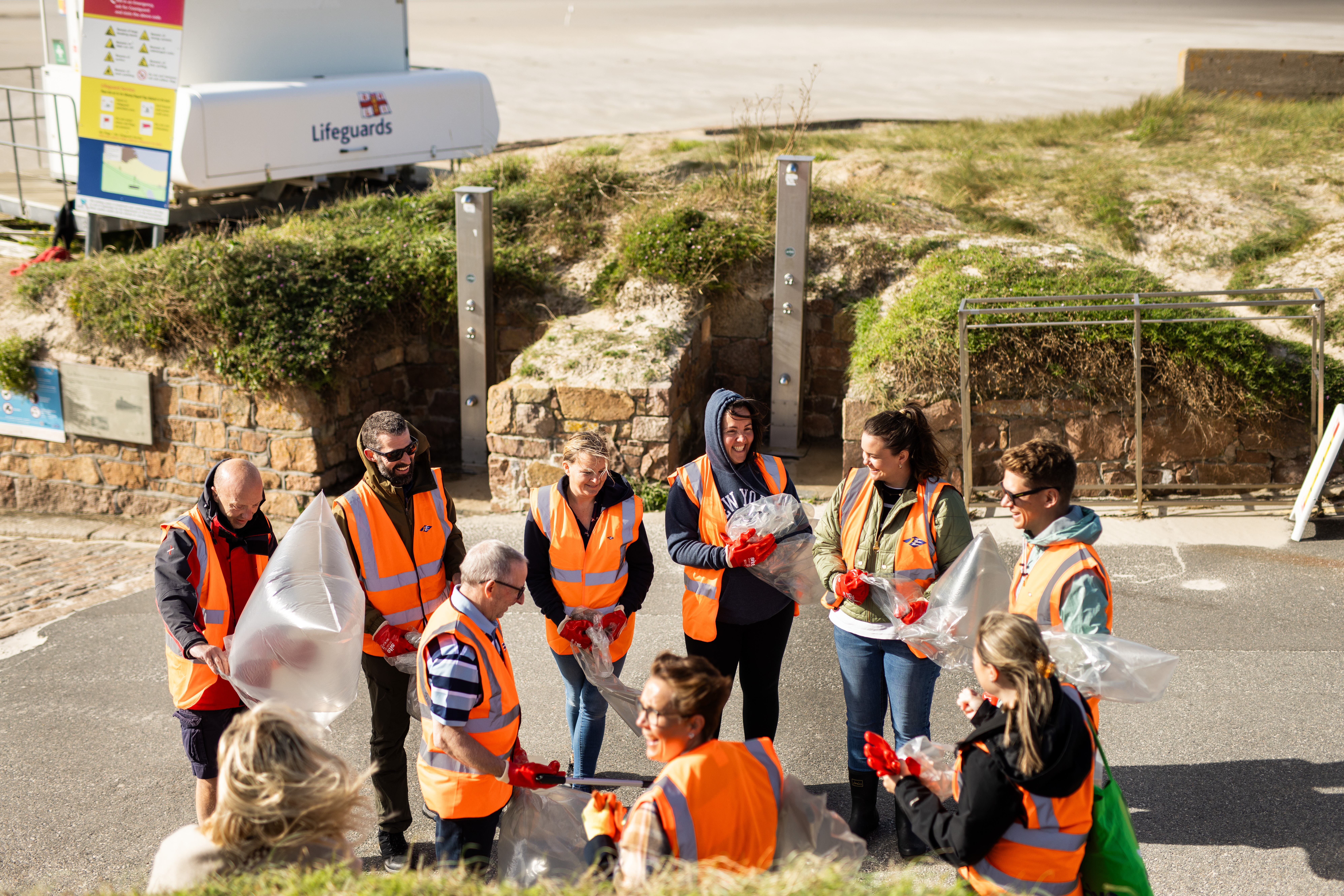 A group of smiling people on a sunny day standing in a beach carpark wearing high vis jackets and holding rubbish bags