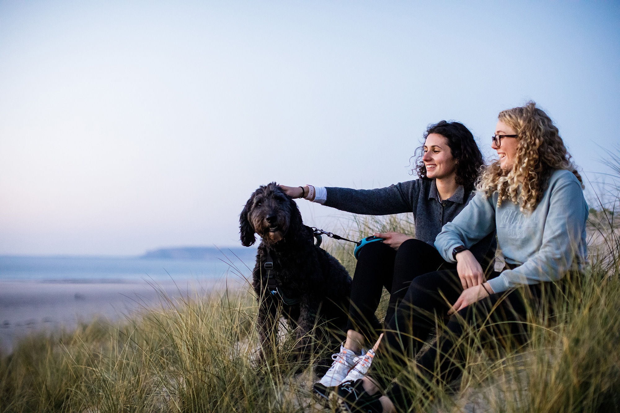 Two female friends sit on the sand dunes at St Ouens, the girl on the left pats a black dog.