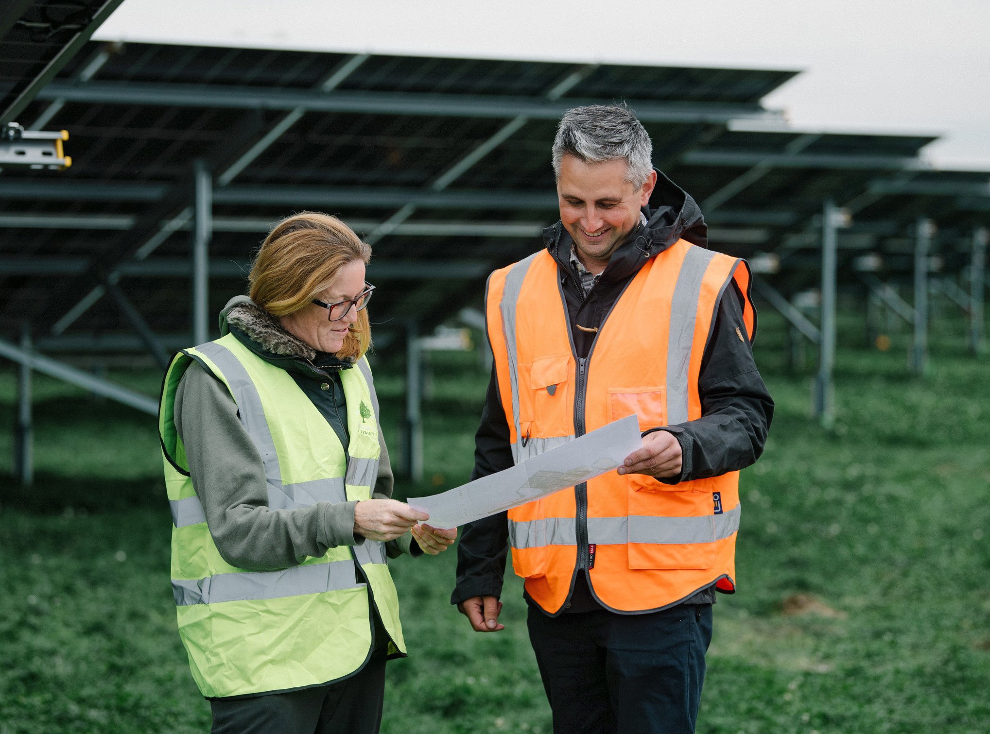 Two people standing in a field with ground mount solar panels behind them, wearing high vis jackets and looking at a piece of paper