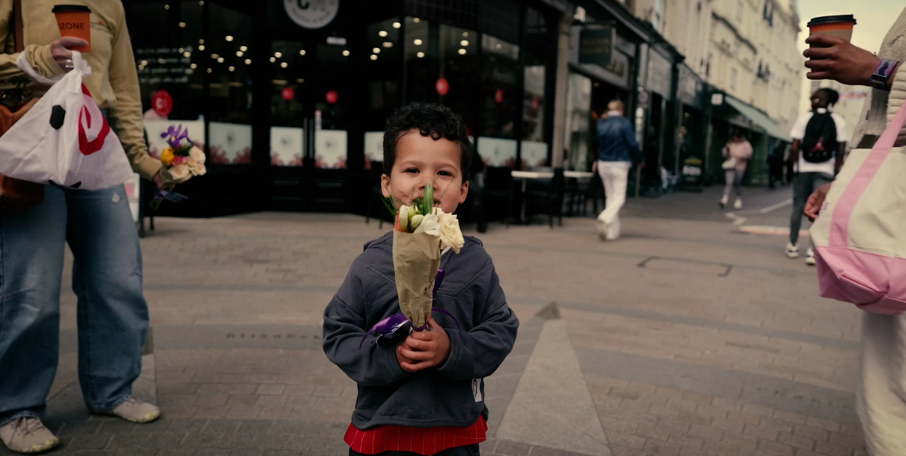 A young boy smiling, holings a small bunch of flowers to his face. Shops and passers by are in the background