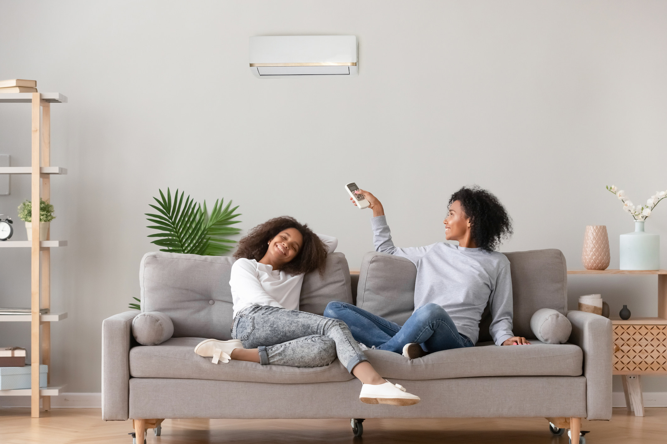 A mother and daughter sit on a couch in a modern home. The mother is operating her air conditioning unit with the remote.