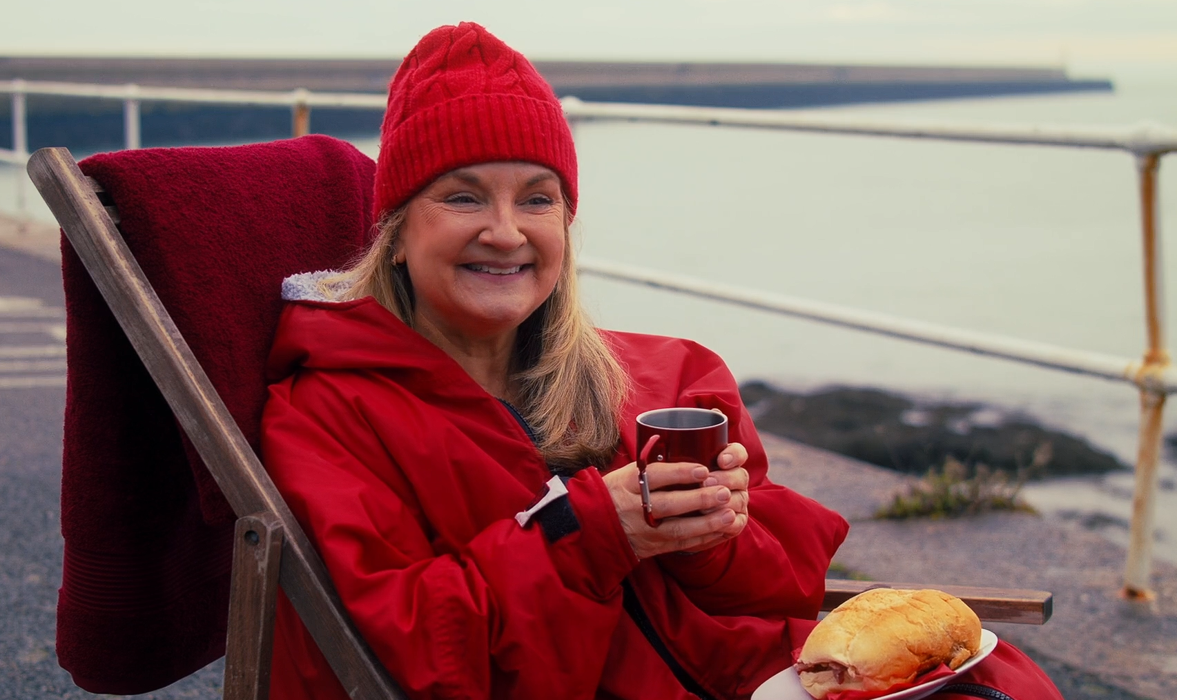Woman in red beanie and jacket seated in a deckchair, holding a red mug and smiling, with a sandwich on a plate; sea and white railing in the background.
