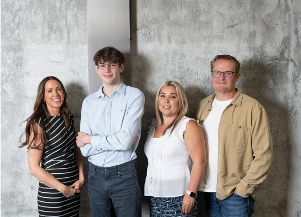 Four people stand together in front of a textured gray wall. A woman in a black and white striped dress stands on the far left, followed by a young man in a light blue button-down shirt, a woman in a white ruffled top, and a man in a tan button-down shirt over a white t-shirt on the far right.
