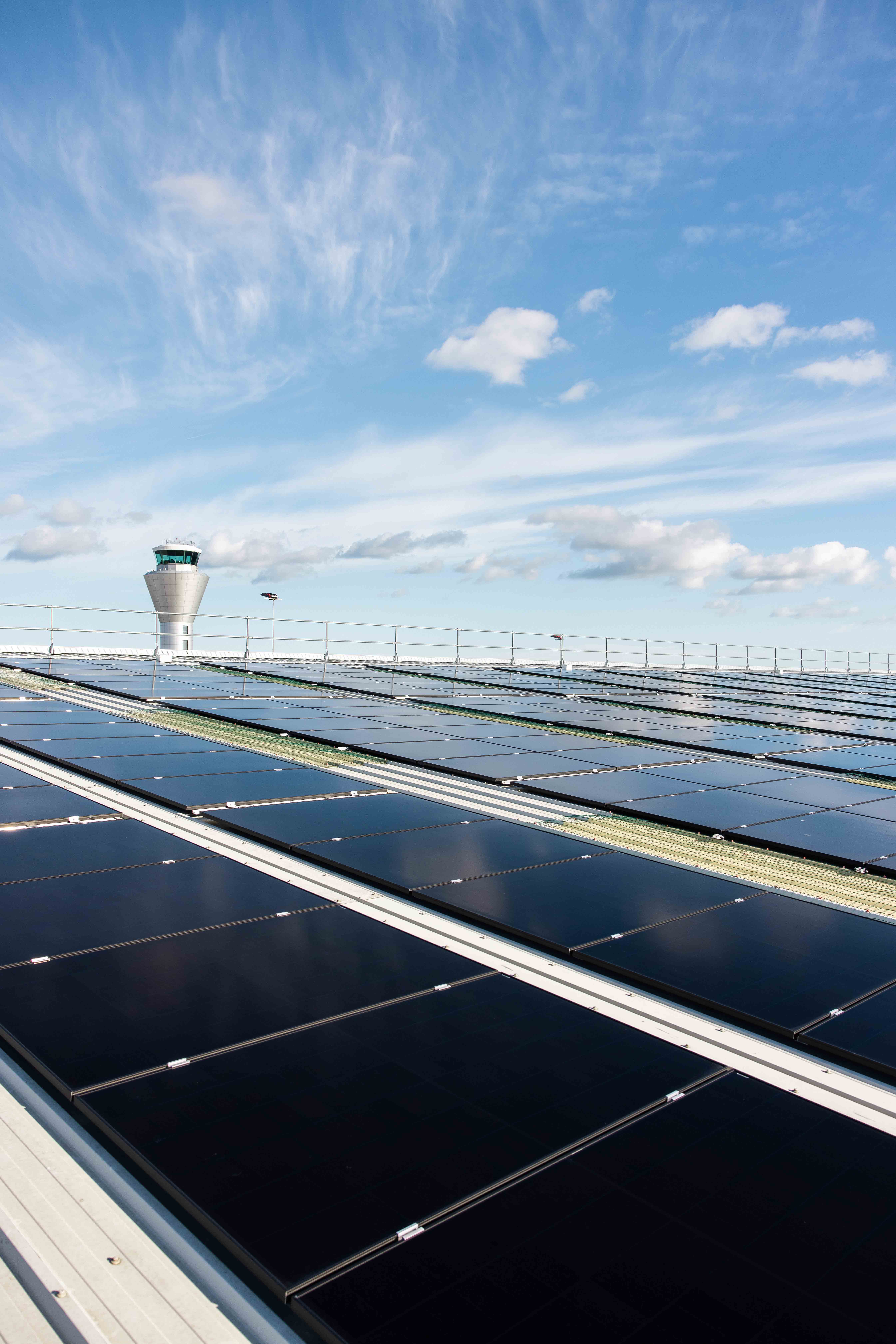 Rows of black solar panels installed on a large rooftop under a bright blue sky with scattered clouds. In the background, an airport control tower is visible, indicating the location near an airport facility.