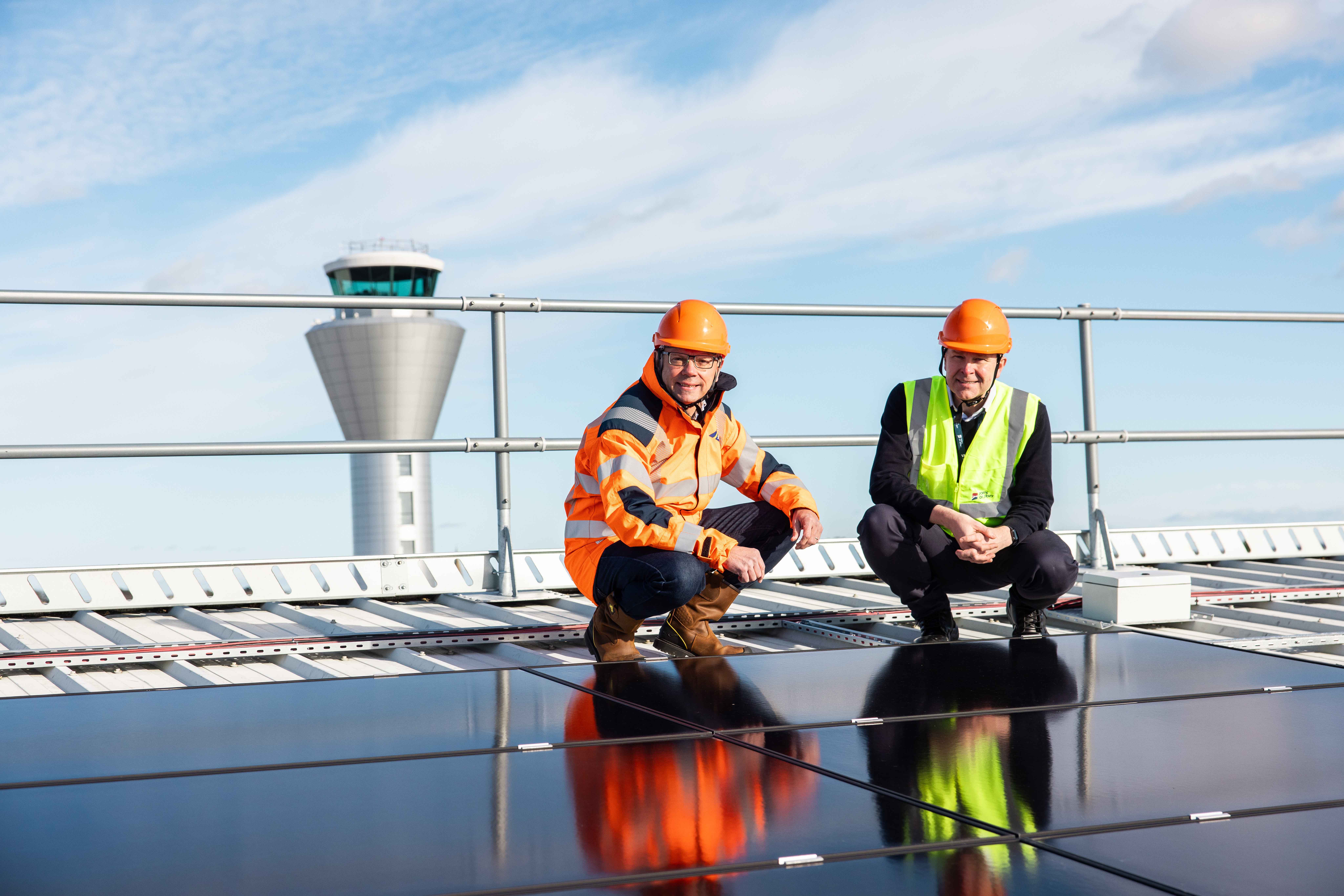 Two people in high vis jackets and hard hats crouched on a roof next to solar panels with the airport control tower in the background
