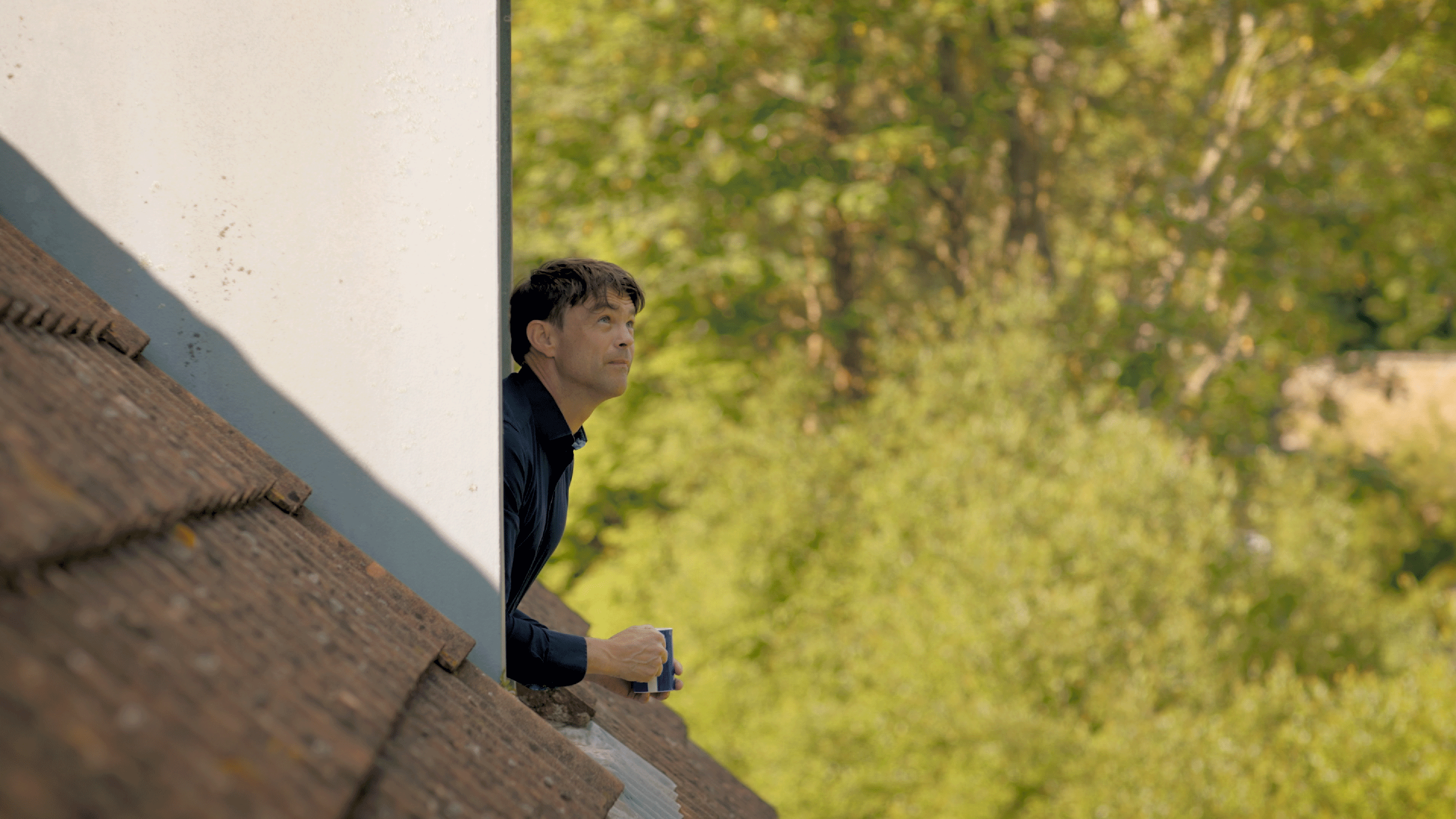 A close up photo of the side of a roof of a home. There's a man in a navy blue top looking out of the dormer window, gazing into the distance. The background of the photo is of trees.