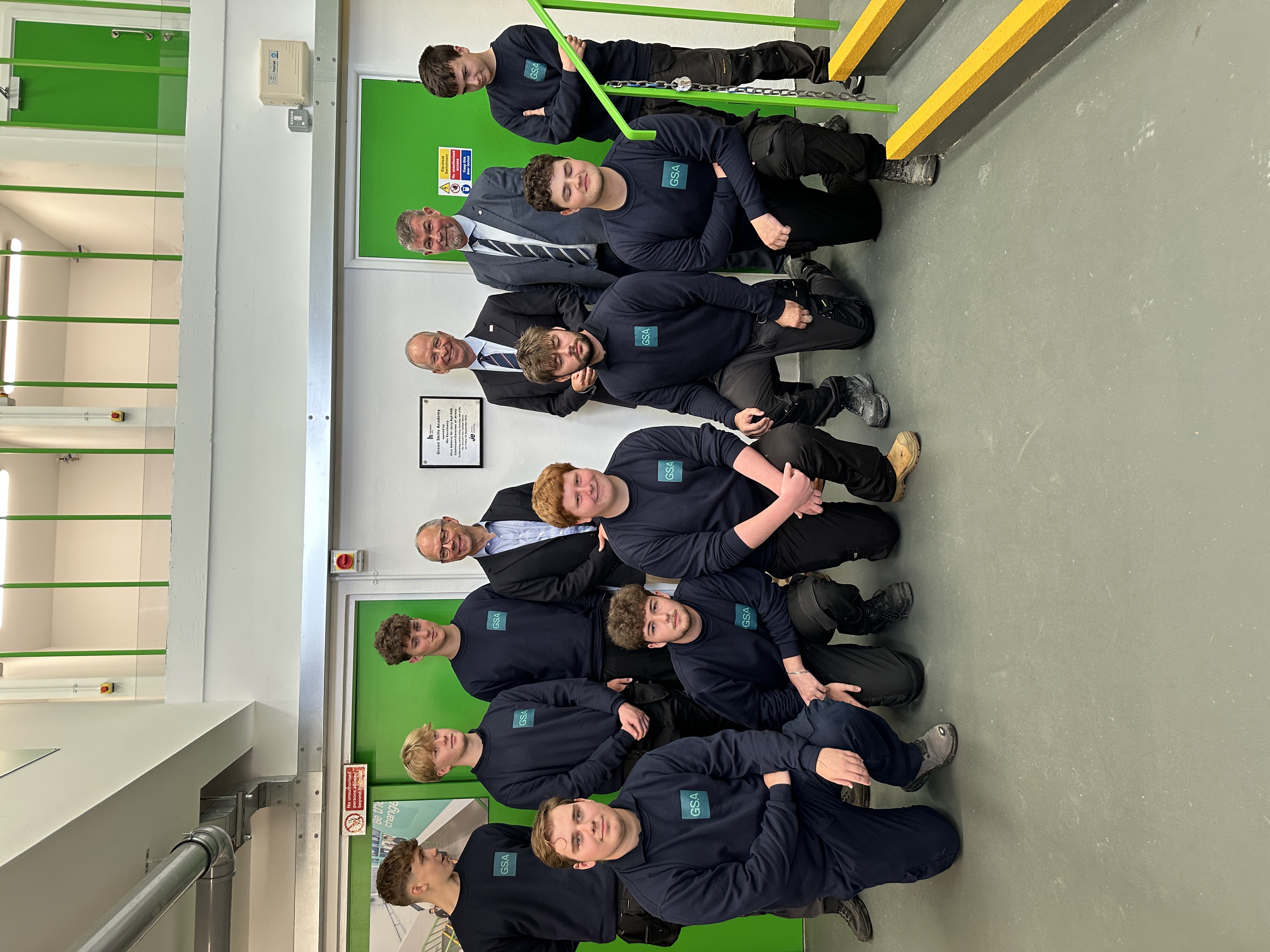 A group of Green Skills Academy apprentices wearing their Green Skills uniform together indoors in front of a plaque on the wall. Standing with them, wearing suits, are Chris Amber - JE CEO, Lieutenant Governor Vice Admiral Sir Jerry Kyd KBE, and Lance Chatfield - Principal of Highlands College. The room has green doors and a white wall.