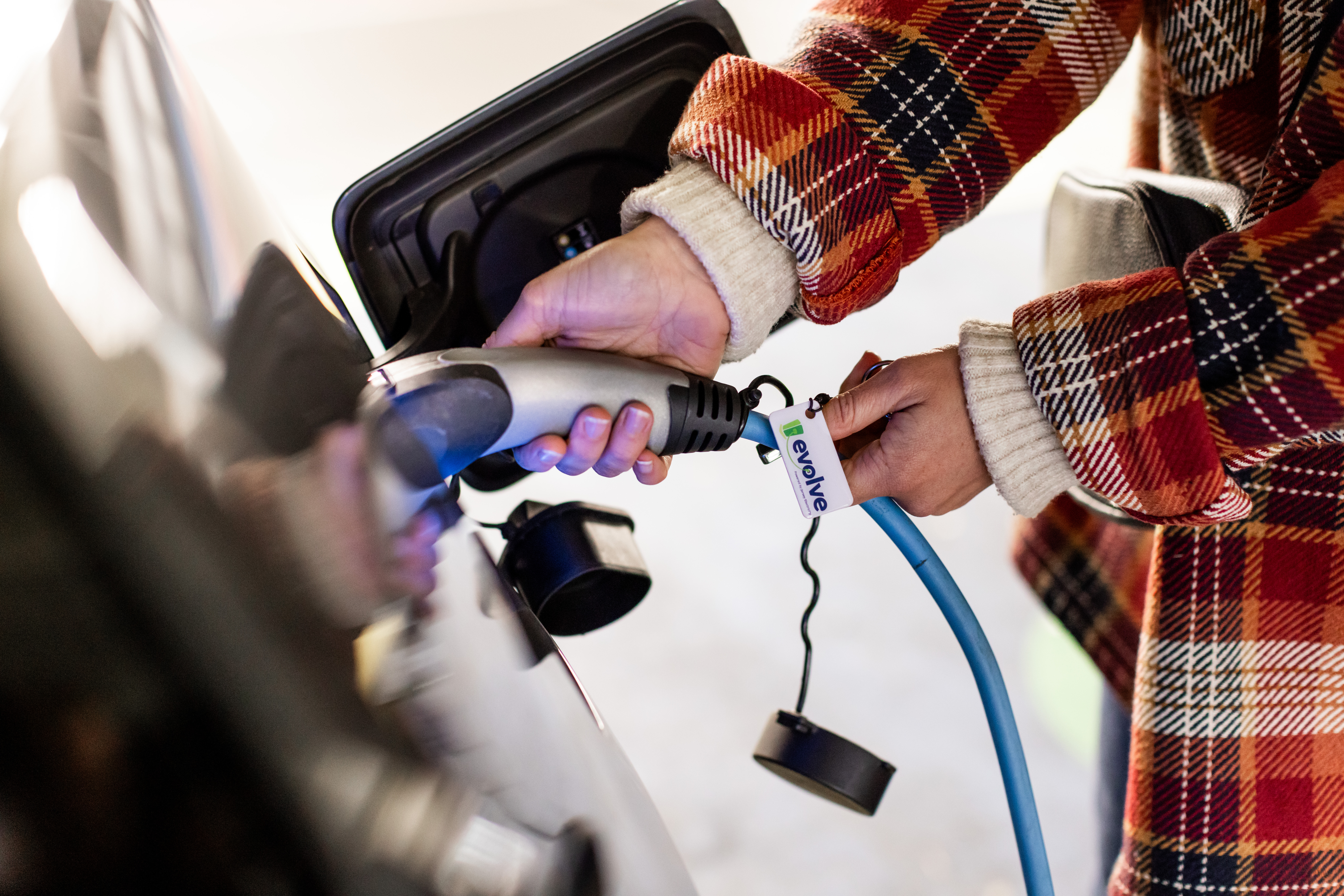 A close up photo of a person in a red checked jacket plugging in a charging cble to their electric vehicle