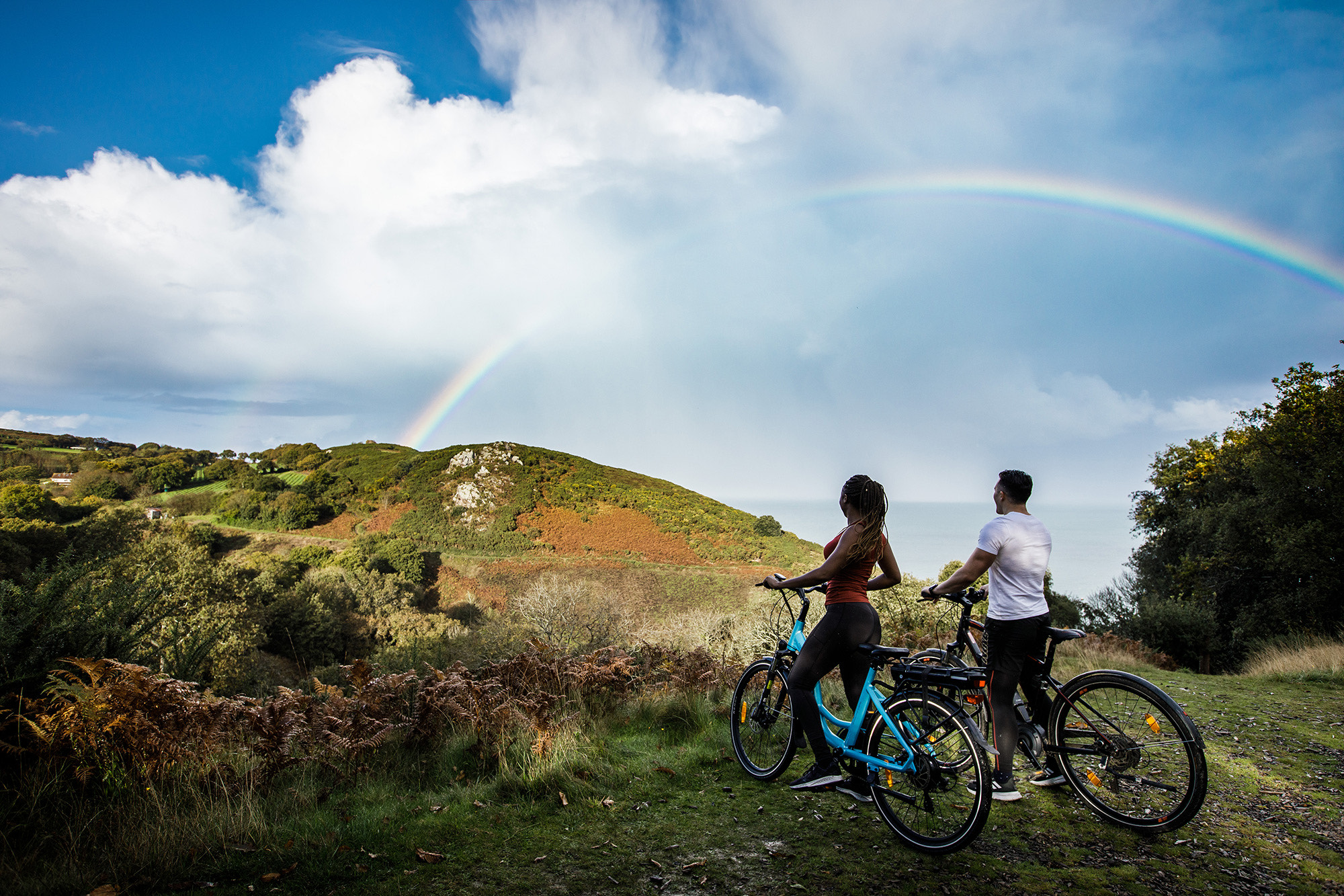A man and a women look at a rainbow while riding electric bicycles.
