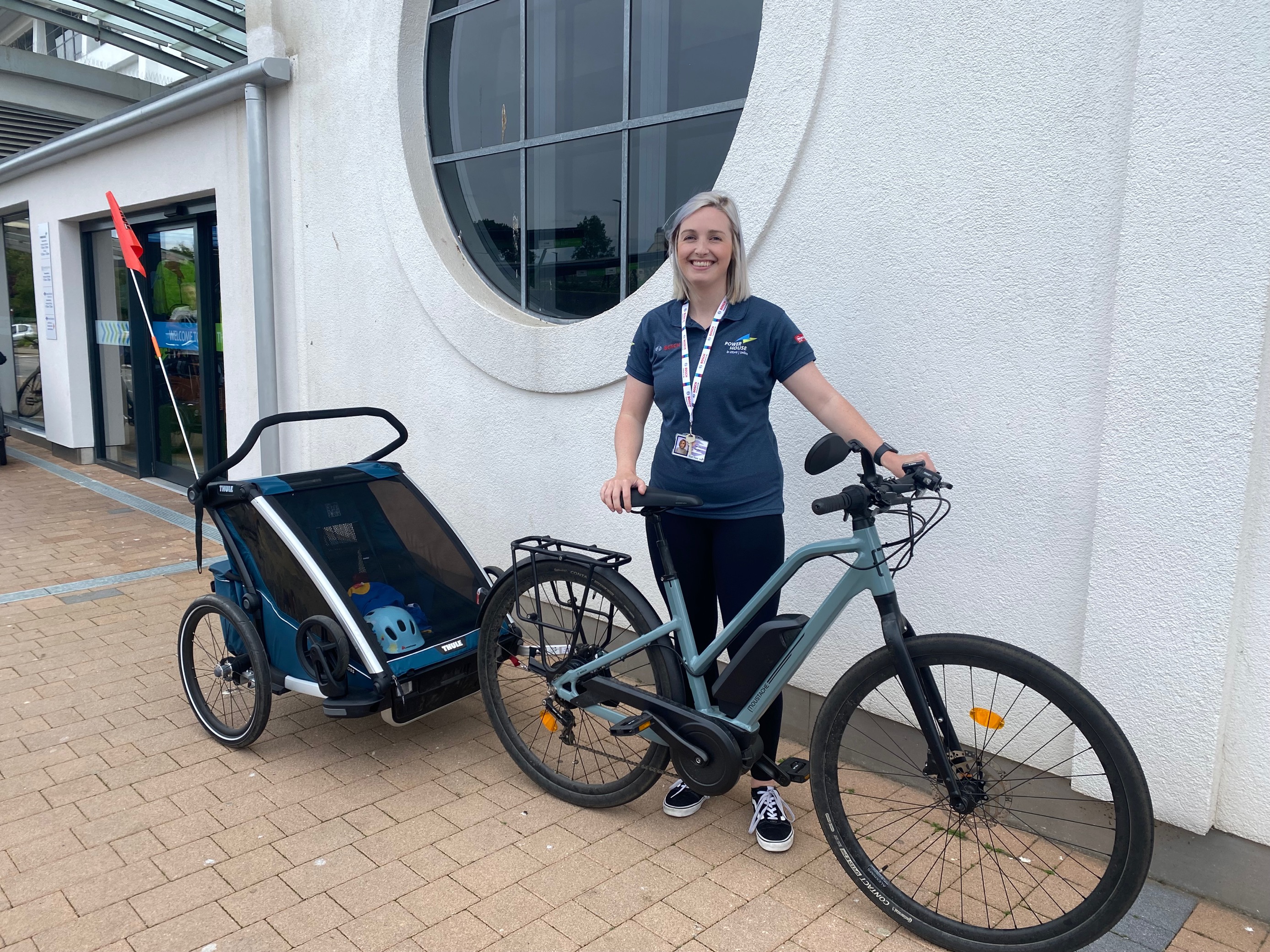 Sarah Naylor, Category Manager at the Powerhouse standing in a blue Powerhouse t-shirt next to her electric bike with Thule Chariot Cross trailer