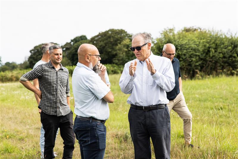 A photo of two men in white shirts and dark trousers deep in discussion. The man on the right is wearing sunglasses. The conversation is taking place in a green field. In the background are people talking and trees.