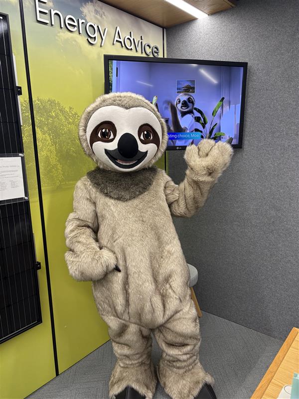 A person in a life size sloth suit, waving. Behind them is a TV screen featuring an animated sloth