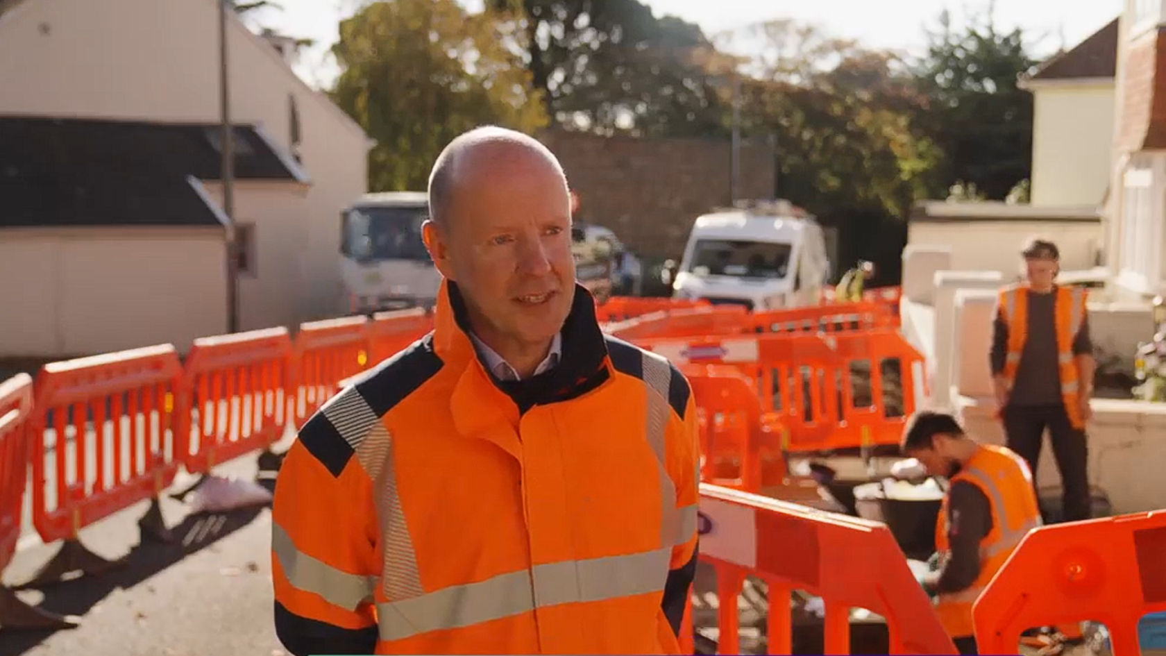 A person wearing orange high vis overalls on a sunny day, standing next next to roadworks